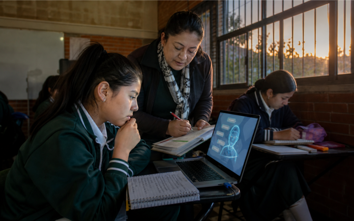 Estudiante y maestra revisan un ejercicio junto a una computadora con un tutor de inteligencia artificial en un aula mexicana.