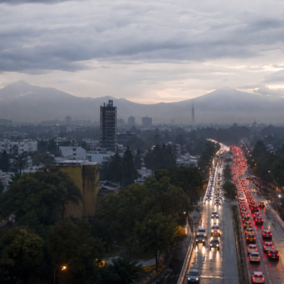Tráfico vehicular en una avenida de la Ciudad de México al amanecer, con pavimento mojado y cielo nublado.