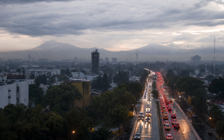 Tráfico vehicular en una avenida de la Ciudad de México al amanecer, con pavimento mojado y cielo nublado.