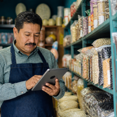 Pequeña tienda con anaqueles etiquetados y una persona registrando existencias en una tableta.