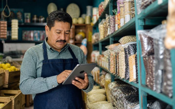 Pequeña tienda con anaqueles etiquetados y una persona registrando existencias en una tableta.
