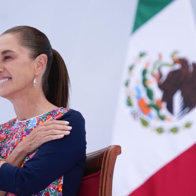 Claudia Sheinbaum sonríe sentada junto a la bandera de México durante el acto.