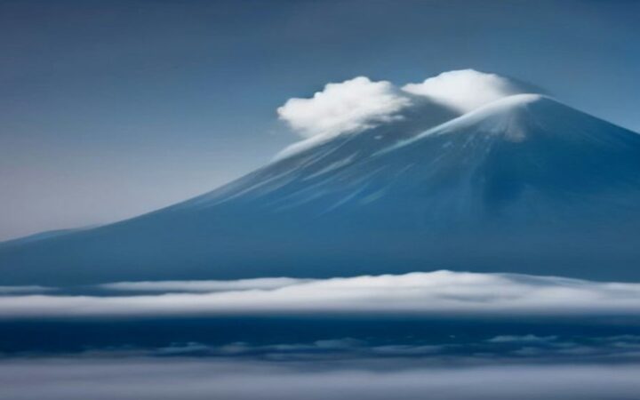 Volcán con cumbre nevada rodeado de nubes y niebla bajo un cielo azul frío al amanecer